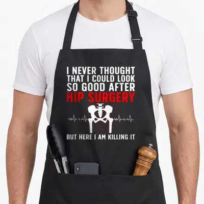 Recovering hip apron - White pelvis on red grid with text I never thought that I could look so good after hip surgery — shown on a black cotton canvas apron against a white background