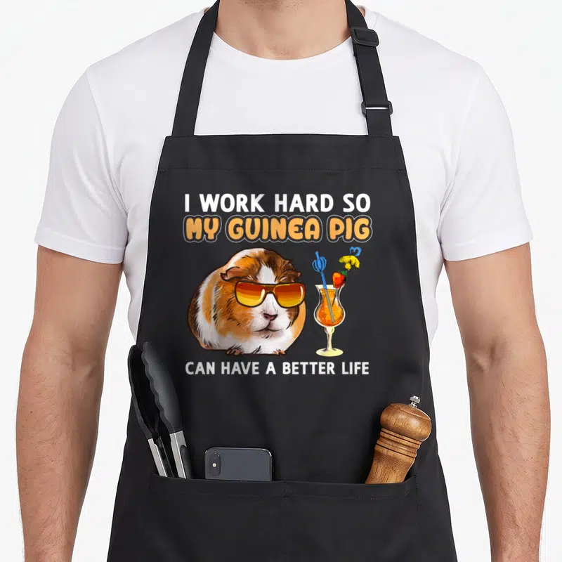 Funny pig crazy guinea man apron - Cartoon guinea pig in sunglasses next to orange cocktail with text about working hard — shown on a black cotton canvas apron against a white background