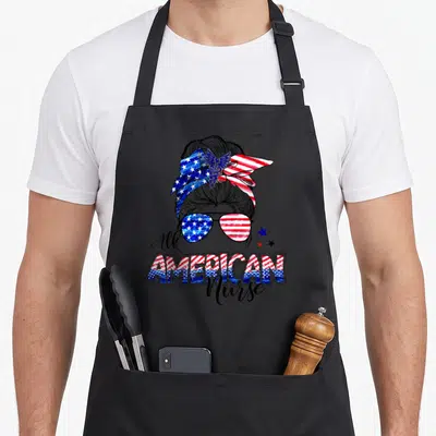 Nurse 4th of july apron - Cartoon nurse head with flag bun, sunglasses, and All AMERICAN Nurse text — shown on a black cotton canvas apron against a white background