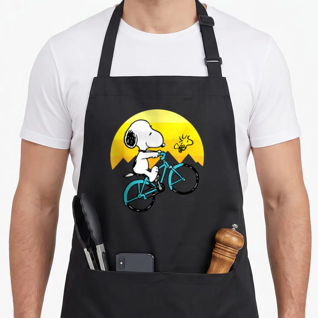 Woodstock biking apron - Snoopy riding blue bike in front of yellow orange striped sun with Woodstock flying nearby — shown on a black cotton canvas apron against a white background