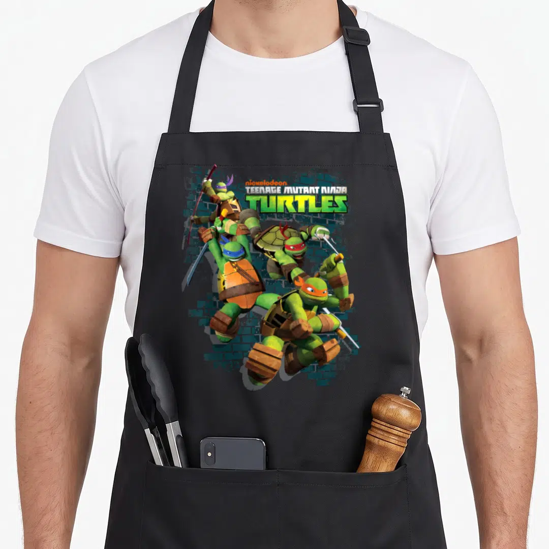 Action teenage cooking aprons - Four turtles leaping forward with logo above brick wall background — shown on a black cotton canvas apron against a white background