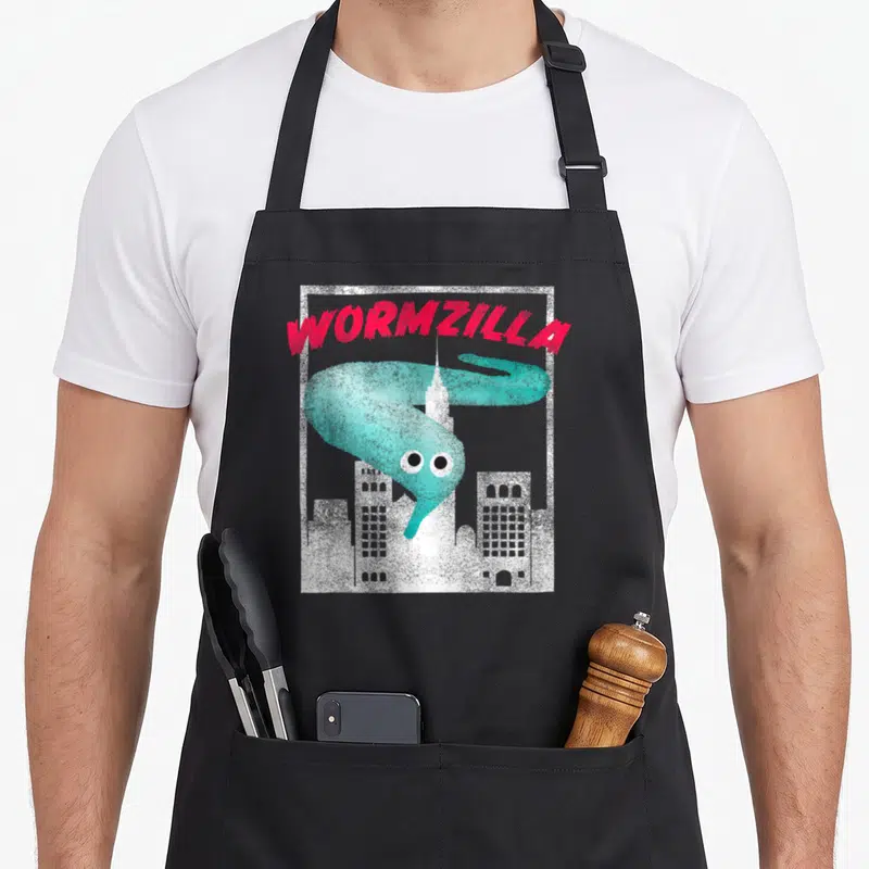 String meme apron - Red WORMZILLA text arching over teal worm and white city skyline inside white frame — shown on a black cotton canvas apron against a white background