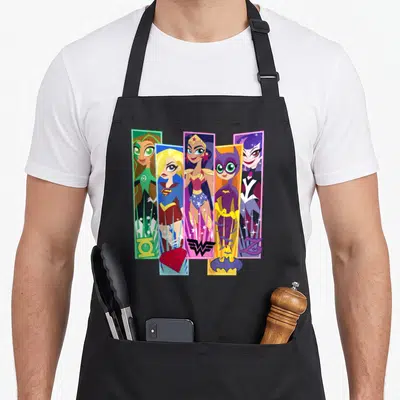 Hero character aprons - Five vertical cartoon hero panels in purple, blue, red, green, yellow with bottom logos — shown on a black cotton canvas apron against a white background