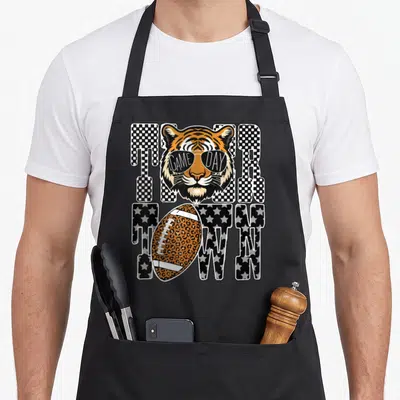 Tiger leopard apron - Cartoon tiger head wearing sunglasses over TIGER TOWN text with leopard print football — shown on a black cotton canvas apron against a white background