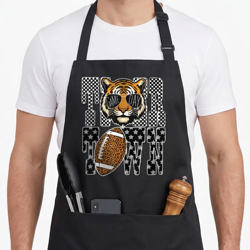 Tiger leopard apron - Cartoon tiger head wearing sunglasses over TIGER TOWN text with leopard print football — shown on a black cotton canvas apron against a white background