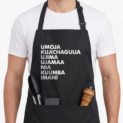 Kwanzaa kwanga apron - Seven white principle names stacked vertically on black fabric — shown on a black cotton canvas apron against a white background