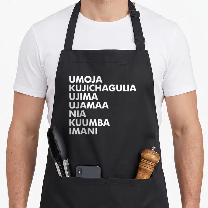 Kwanzaa kwanga apron - Seven white principle names stacked vertically on black fabric — shown on a black cotton canvas apron against a white background