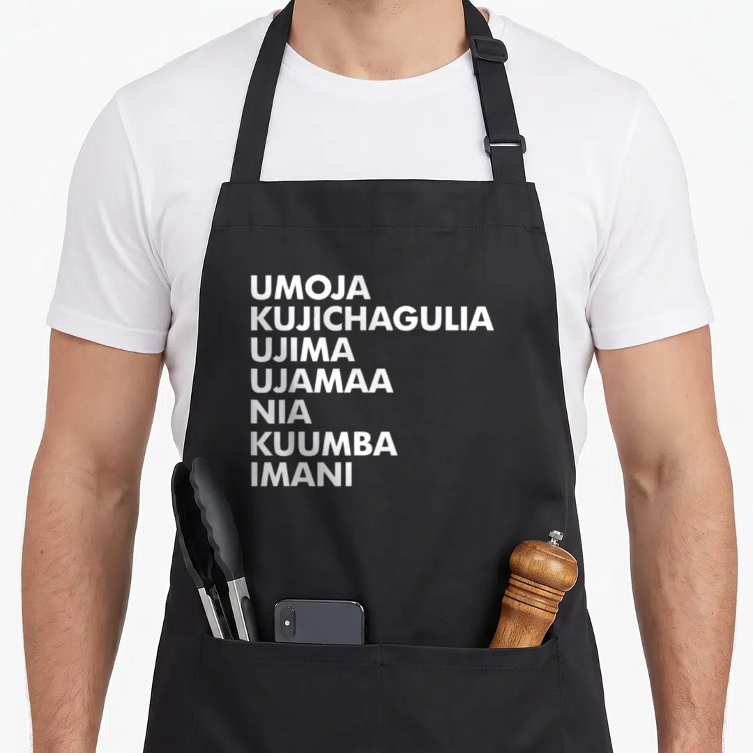 Kwanzaa kwanga apron - Seven white principle names stacked vertically on black fabric — shown on a black cotton canvas apron against a white background