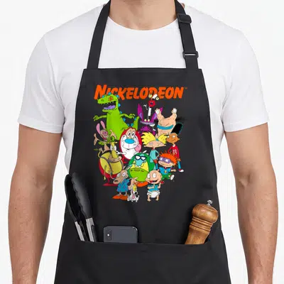 90s character aprons - NICKELODEON text above a cluster of cartoon characters in orange green yellow purple and red — shown on a black cotton canvas apron against a white background