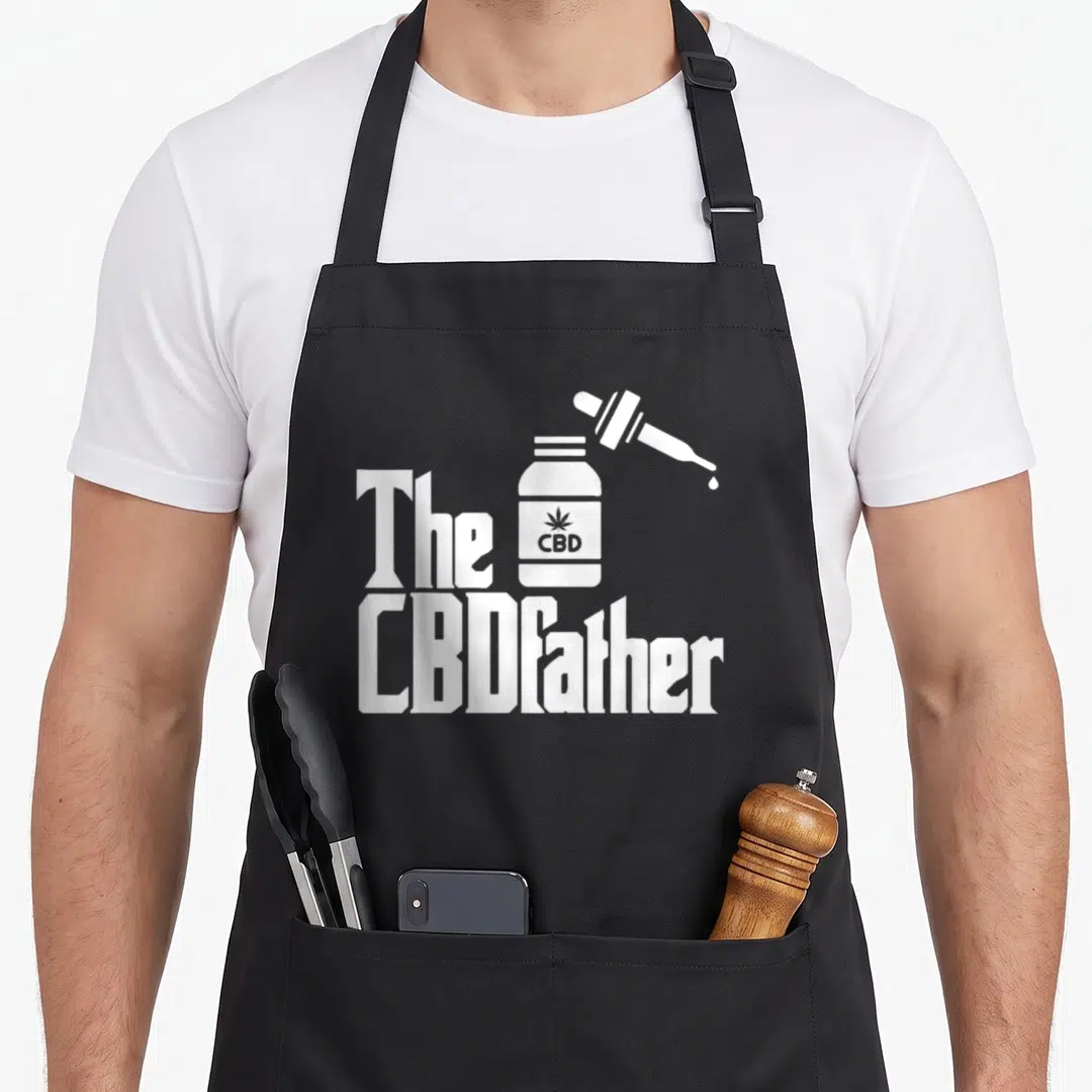 Father hemp apron - White cartoon CBD bottle and dropper with The CBDFather text on black background — shown on a black cotton canvas apron against a white background