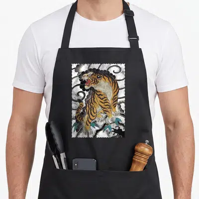 Traditional tattoo apron - Realistic orange tiger standing on rocks with green leaves and grey clouds inside a stamp — shown on a black cotton canvas apron against a white background