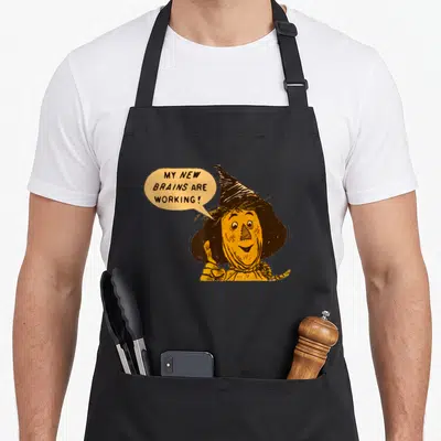Tin lion dorothy apron - Cartoon scarecrow giving thumbs up with speech bubble text MY NEW BRAINS ARE WORKING — shown on a black cotton canvas apron against a white background