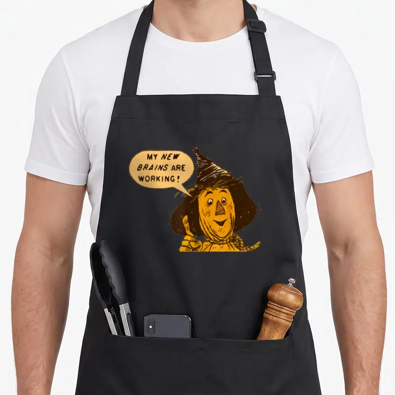 Tin lion dorothy apron - Cartoon scarecrow giving thumbs up with speech bubble text MY NEW BRAINS ARE WORKING — shown on a black cotton canvas apron against a white background