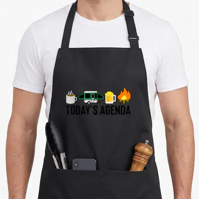 Today happy camper apron - TODAY'S AGENDA text below four icons: mug, green trailer, beer, fire in cartoon style — shown on a black cotton canvas apron against a white background