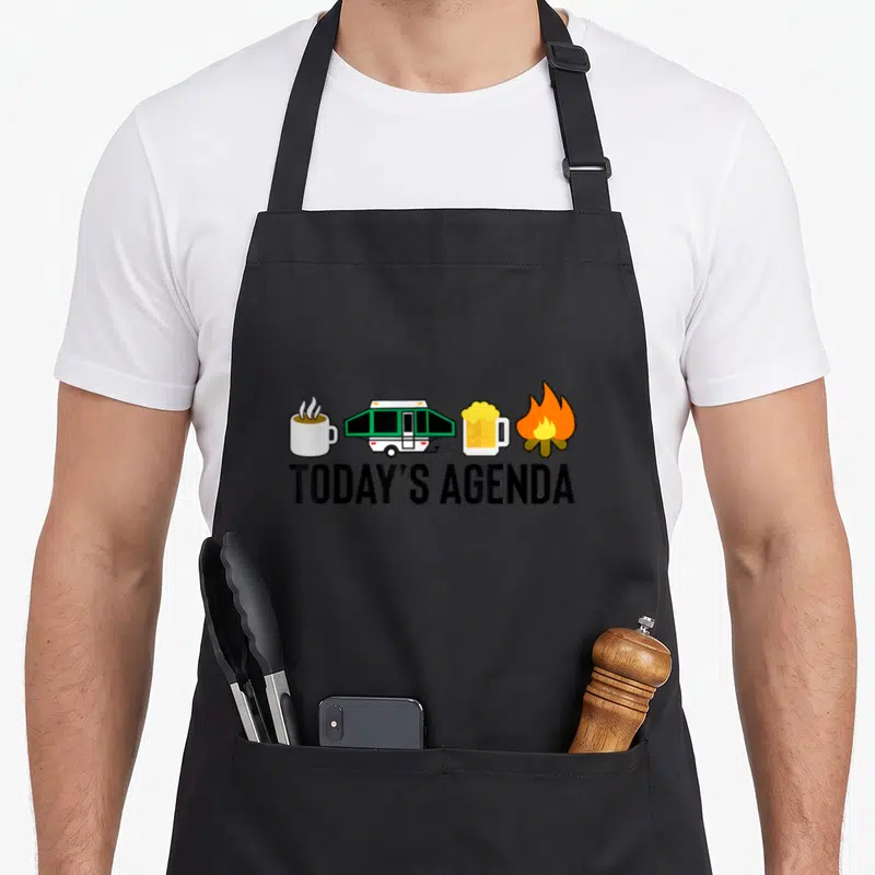 Today happy camper apron - TODAY'S AGENDA text below four icons: mug, green trailer, beer, fire in cartoon style — shown on a black cotton canvas apron against a white background