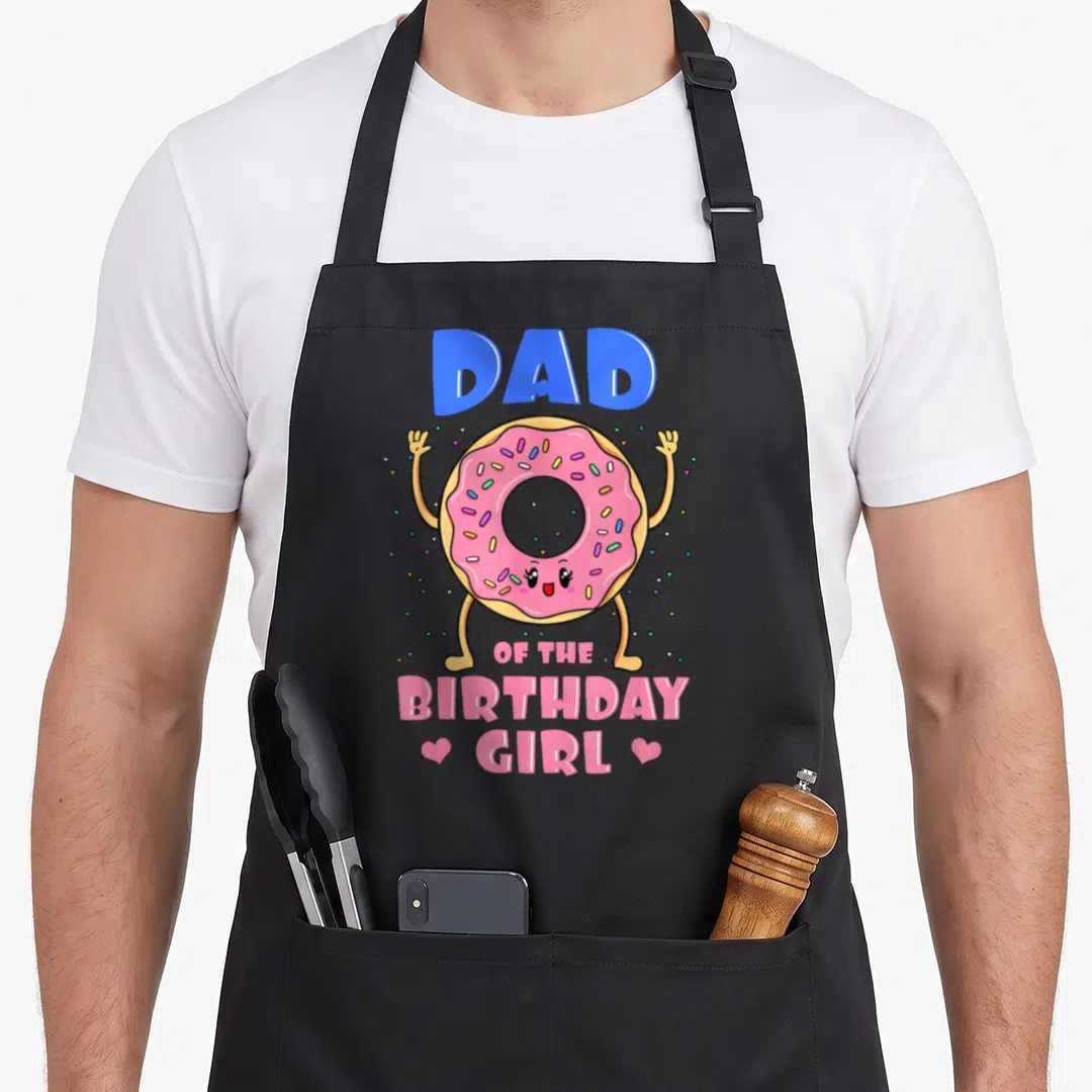 Donut grill daddy apron - Pink frosted donut character with blue DAD text above and pink OF THE BIRTHDAY GIRL below — shown on a black cotton canvas apron against a white background