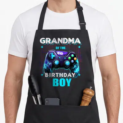 Grandma video game apron - Retro purple game controller with GRANDMA OF THE BIRTHDAY BOY text and pink grid background — shown on a black cotton canvas apron against a white background