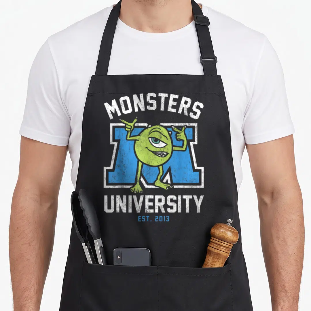 Monster collegiate aprons - Green one-eyed monster stands before blue M with white MONSTERS UNIVERSITY text and EST — shown on a black cotton canvas apron against a white background