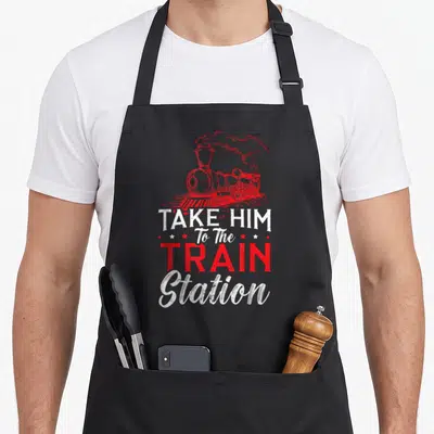 Railway train apron - Red steam train illustration with distressed white and red text reading Take Him To The Train — shown on a black cotton canvas apron against a white background