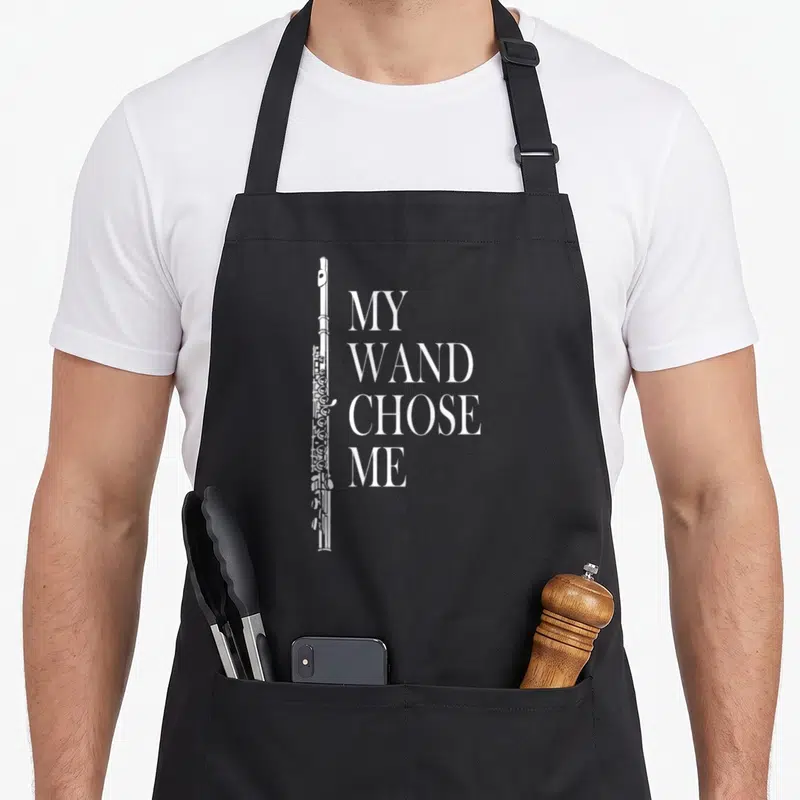 Wand flute apron - Vertical white flute beside stacked text reading My Wand Chose Me in serif font — shown on a black cotton canvas apron against a white background