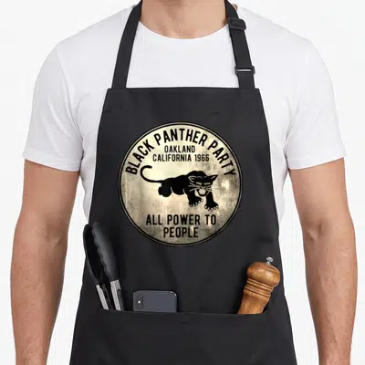 Funny oakland apron - Circular beige emblem with black panther and text BLACK PANTHER PARTY OAKLAND 1966 ALL POWER — shown on a black cotton canvas apron against a white background