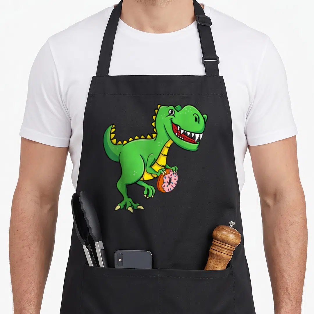 Donut dinosaur apron - Green cartoon T-Rex holding a pink frosted doughnut with multicolored sprinkles — shown on a black cotton canvas apron against a white background
