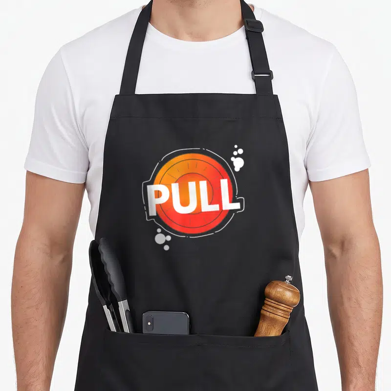Trap shooting clay apron - Cartoon orange and red button with white PULL text and scattered bubbles — shown on a black cotton canvas apron against a white background