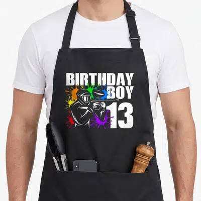 Paintball 13th birthday apron - Cartoon paintball player silhouette with BIRTHDAY BOY 13 text and colorful splatters — shown on a black cotton canvas apron against a white background