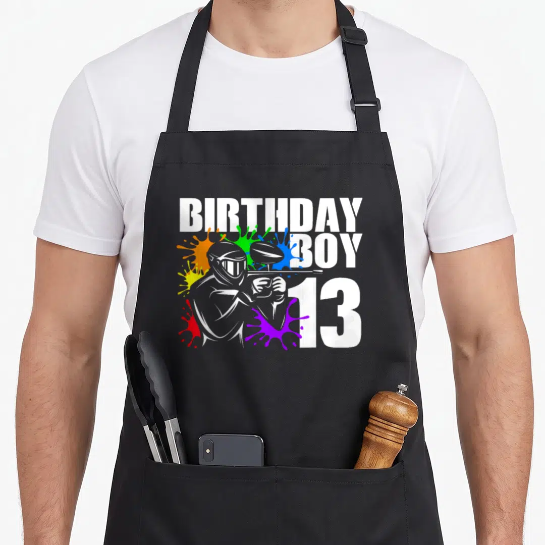 Paintball 13th birthday apron - Cartoon paintball player silhouette with BIRTHDAY BOY 13 text and colorful splatters — shown on a black cotton canvas apron against a white background
