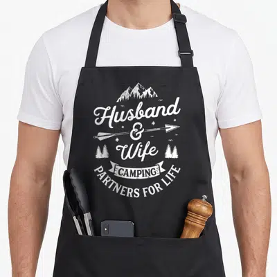 Wife camping apron - White retro text reading Husband Wife Camping Partners For Life with mountains and arrow on black — shown on a black cotton canvas apron against a white background