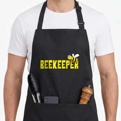 Keeper bee apron - Cartoon bee perched on yellow BEEKEEPER text with black stripes and white wings — shown on a black cotton canvas apron against a white background