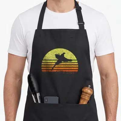 Pheasant bird apron - Grey bird silhouette flying across a yellow and orange striped retro sun graphic — shown on a black cotton canvas apron against a white background