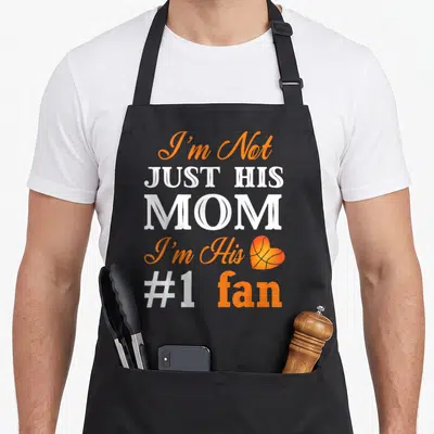 Fan basketball mom apron - I'm Not Just His Mom text with orange basketball heart graphic — shown on a black cotton canvas apron against a white background