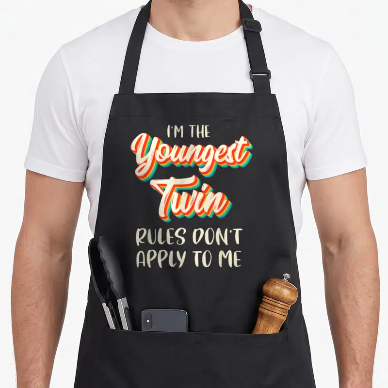 Youngest birthday apron - Stacked colorful text reading I'm the Youngest Twin Rules Don't Apply To Me — shown on a black cotton canvas apron against a white background