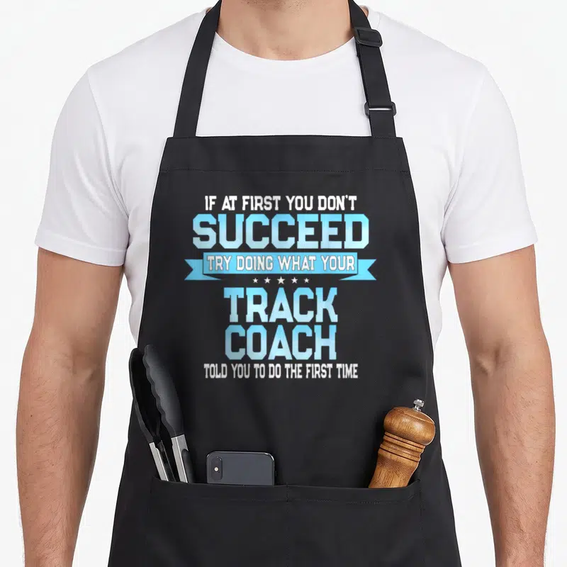 Coach track apron - White and light blue text reading IF AT FIRST YOU DON'T SUCCEED inside a ribbon with five stars — shown on a black cotton canvas apron against a white background