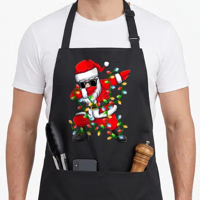Dabbing xmas apron - Cartoon Santa in red and black doing the dab pose wrapped in multicolored Christmas lights — shown on a black cotton canvas apron against a white background