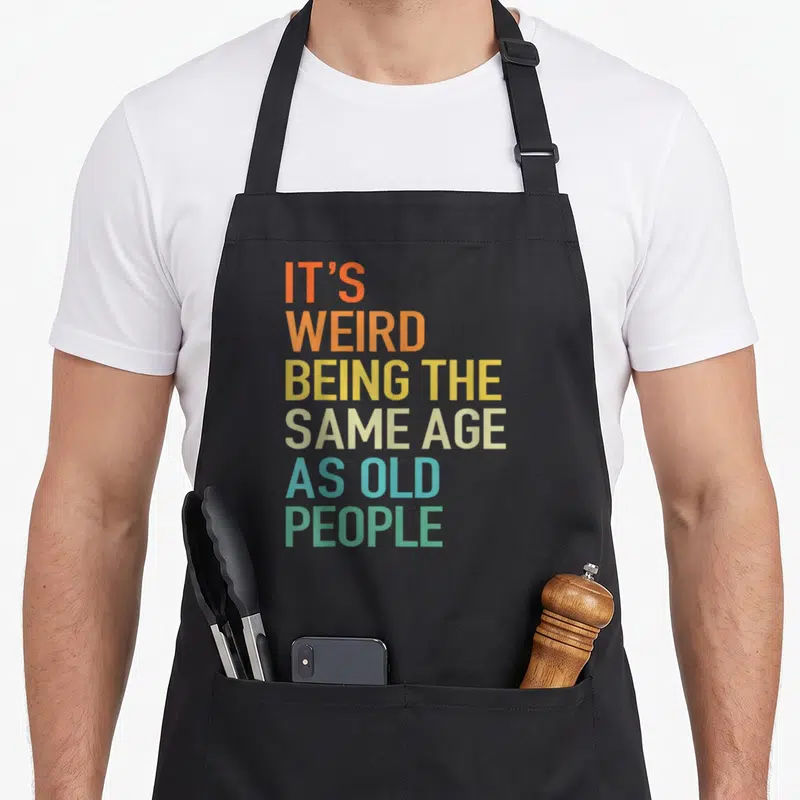 Husband weird aprons - Stacked colorful text reading It's Weird Being The Same Age As Old People — shown on a black cotton canvas apron against a white background