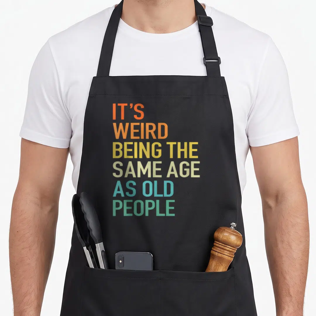 Husband weird aprons - Stacked colorful text reading It's Weird Being The Same Age As Old People — shown on a black cotton canvas apron against a white background