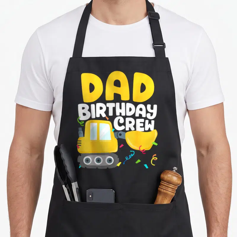 Birthday crew truck apron - Yellow cartoon bulldozer with stacked DAD BIRTHDAY CREW text and confetti — shown on a black cotton canvas apron against a white background