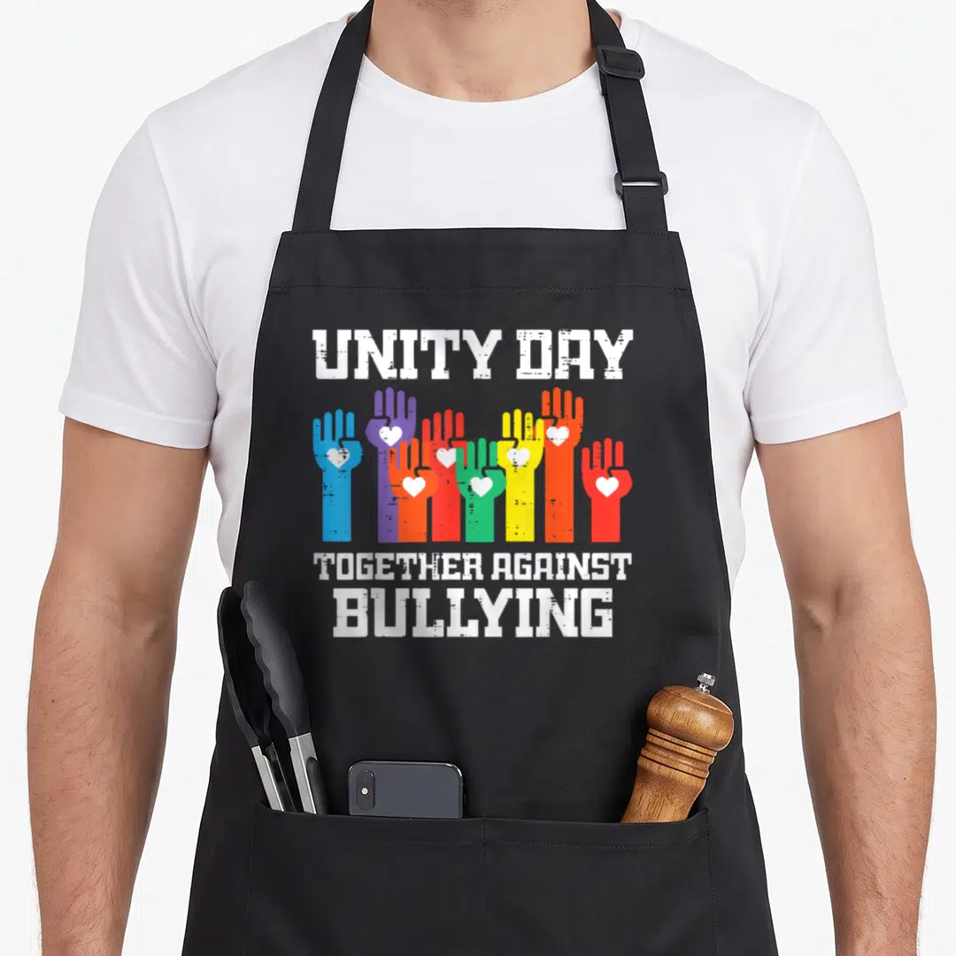 Against unity day apron - Seven multicolored hands with white hearts and distressed white text reading unity day — shown on a black cotton canvas apron against a white background