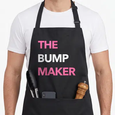 Pregnancy makers apron - Pink and white stacked text reading The Bump Maker — shown on a black cotton canvas apron against a white background