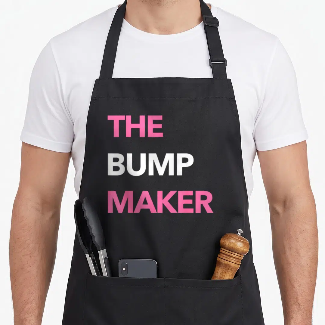 Pregnancy makers apron - Pink and white stacked text reading The Bump Maker — shown on a black cotton canvas apron against a white background
