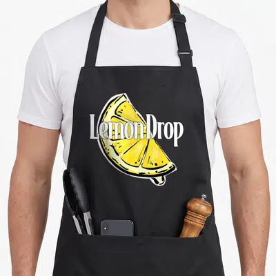 Funny drop for love and lemons apron - LemonDrop text over a yellow cartoon lemon slice with black outlines — shown on a black cotton canvas apron against a white background