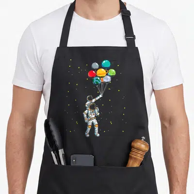 Universe balloon apron - Two cartoon astronauts holding a bunch of colorful planet balloons against a starry background — shown on a black cotton canvas apron against a white background
