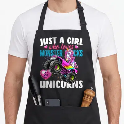 Monster unicorn truck apron - Pink monster truck driven by a unicorn with stacked text Just a Girl who loves monster — shown on a black cotton canvas apron against a white background