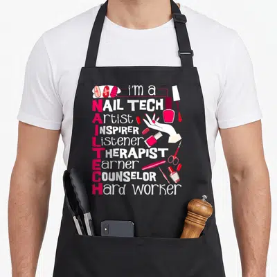 Artist studio manicurist apron - Stacked text listing nail tech roles with vertical pink NAIL TECH acronym — shown on a black cotton canvas apron against a white background