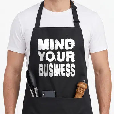 Gossip business apron - White distressed text reading Mind Your Business stacked in three lines — shown on a black cotton canvas apron against a white background