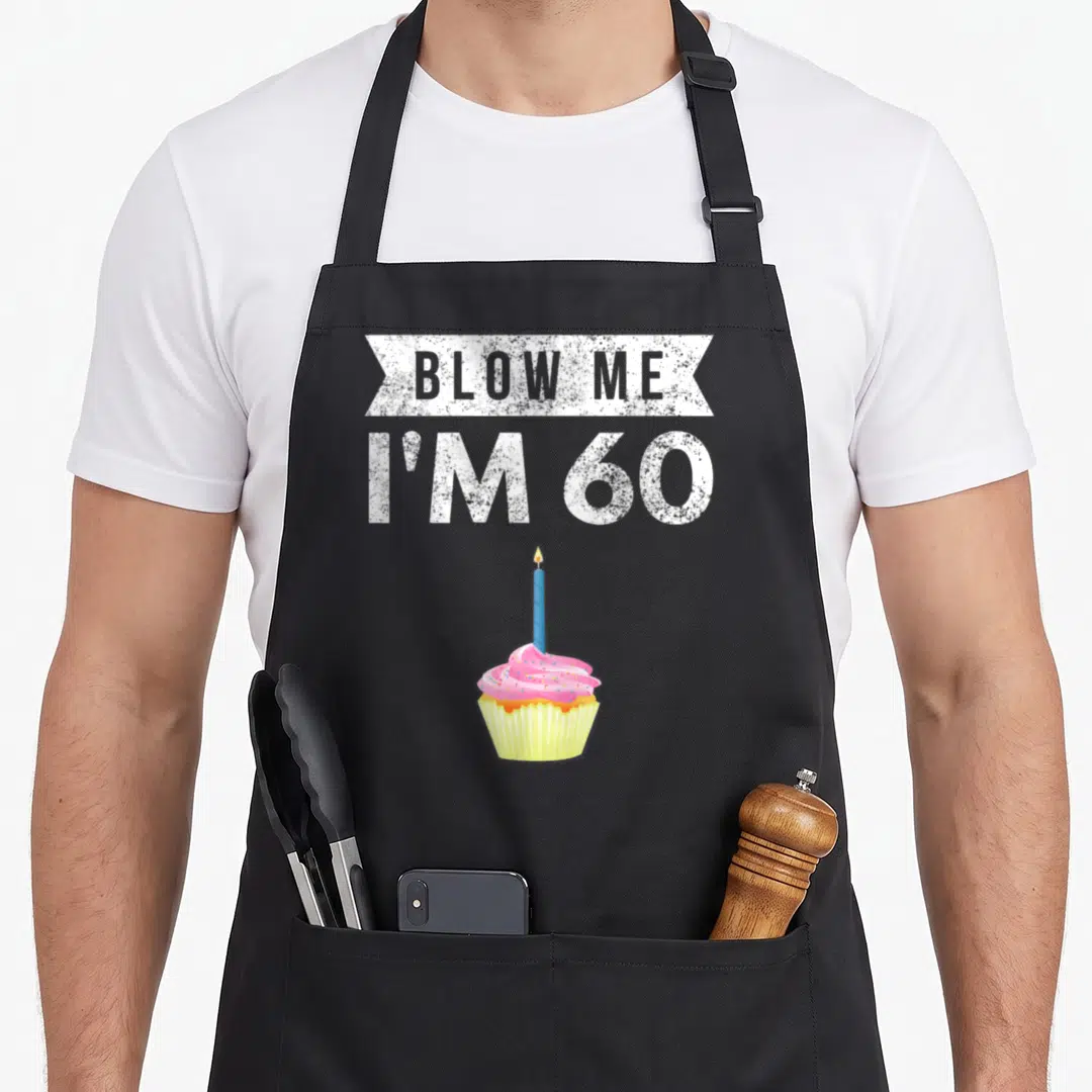Blow 60th birthday apron - Cartoon cupcake with lit candle under BLOW ME I'M 60 text banner — shown on a black cotton canvas apron against a white background