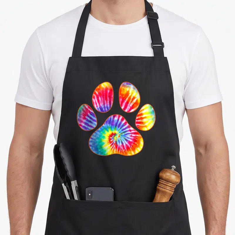 Tie custom pet apron - Tie-dye paw print with orange outline in red yellow green blue and purple — shown on a black cotton canvas apron against a white background