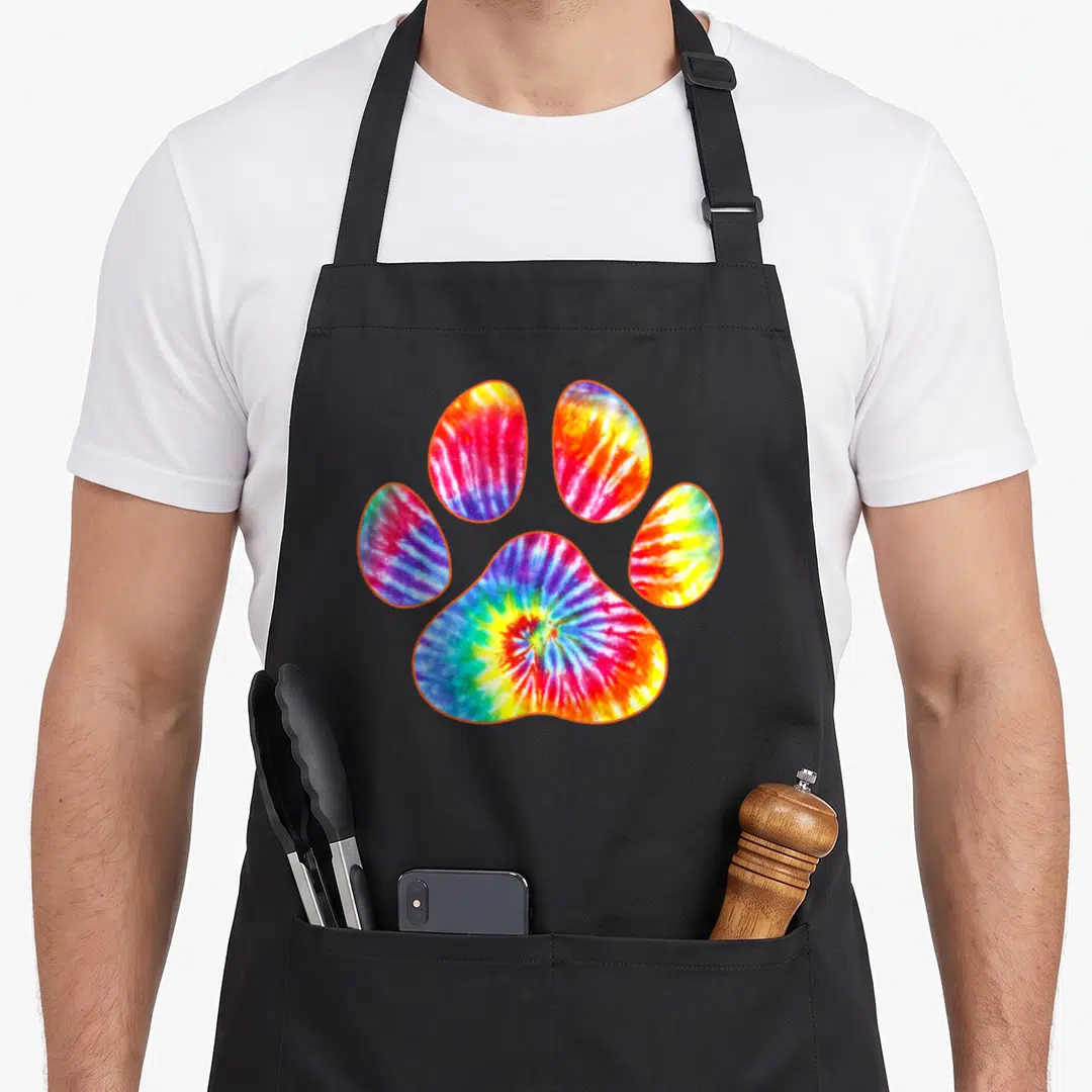 Tie custom pet apron - Tie-dye paw print with orange outline in red yellow green blue and purple — shown on a black cotton canvas apron against a white background
